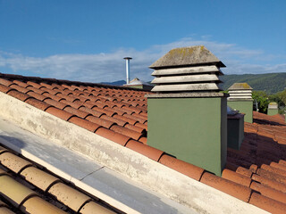 Modern stainless steel oven chimney and brick chimneys on a red tile roof under blue sky with...