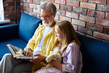 senior man show photo album to granddaughter, they sit on sofa at home, have rest and talk