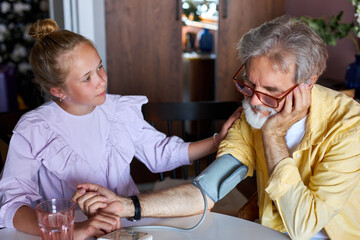 little caucasian child girl take care of sick granddad, measure blood pressure using a tonometer. they sit at table at home