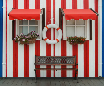 Duas janelas com uma boia no meio e um banco de jardim, toldes das janelas abertos e vasos decorativos pendurados, parede &aacute;s riscas