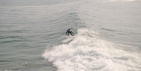 Surfista a surfar de pé em cima prancha na crista de uma onda do mar  com um fato de neoprene vestido ao fim da tarde