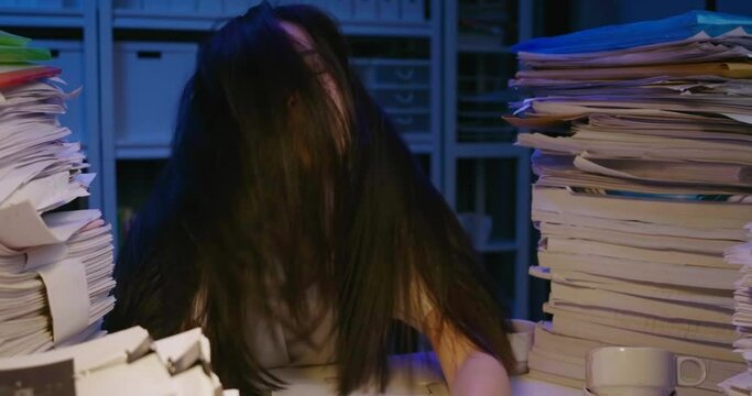 Unhappy young asian student woman is sitting at desk cover with stack of paperwork and mess of her hair. Alone teen girl exhausted, tired and sighting in frustration while studying hard at late night.