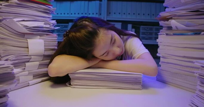 Tired young asian student woman lay down on desk cover with stack of paperwork. Alone teen girl exhausted and sleep on pile of paper sheet while studying hard overload at late night. Dolly shot