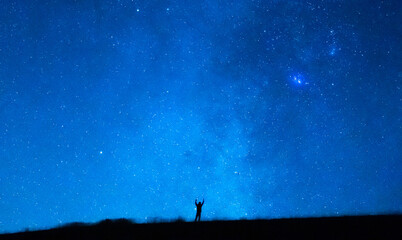 Obraz na płótnie Canvas Person raising arms under the blue starry sky at night. Silhouette of a person observing the immensity of the universe, the stars, the constellations and the planets.
