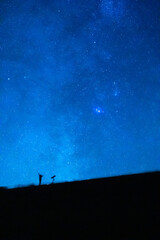 Person with a telescope raising his arms under the blue starry sky at night. Silhouette of a person observing the immensity of the universe.
