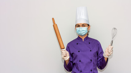 Naklejka premium Woman chef with surgical mask and gloves for coronavirus prevention looks at the camera and holds whisk and rolling pin in her hands.