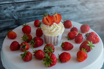 Delicious cupcake in the center with white cream decorated with ripe strawberries on a gray blue background and scattered strawberries.