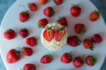 Delicious cupcake in the center with white cream decorated with ripe strawberries on a gray blue background and scattered strawberries.