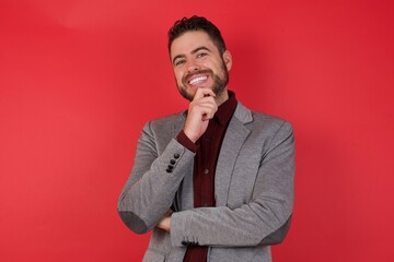 Young caucasian businessman wearing casual clothes standing over isolated red wall Optimistic keeps hands partly crossed and hand under chin, looks at camera with pleasure. Happy emotions concept.