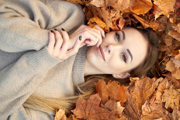 Pretty young girl with charming smile liying in the autumn leaves and enjoying it, holding face and...