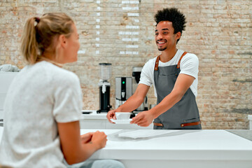 Attractive African male professional barista in apron serves a cup of coffee to a client and smiles.