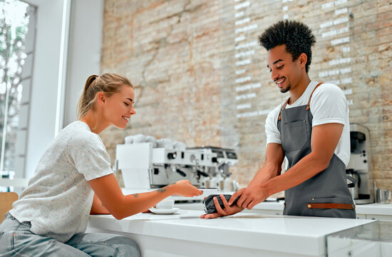 Female Customer Making Payment Through Mobile Phone At Counter In Cafe With Young Man. Barista Holding Credit Card Reading Machine In Front Of Female Costumer With Cell Phone.