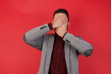 Young caucasian businessman wearing casual clothes standing over isolated red background Covering eyes and mouth with hands, surprised and shocked. Hiding emotions.