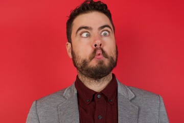 Young caucasian businessman wearing casual clothes standing over isolated red background making fish face with lips, crazy and comical gesture. Funny expression.