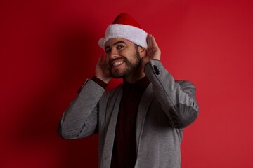 Young caucasian businessman wearing casual clothes and Santa hat standing over isolated red wall Trying to hear both hands on ear gesture, curious for gossip. Hearing problem, deaf