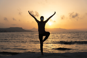 Fototapeta premium Silhouette, young woman with protective surgical face mask performs yoga stretching exercises at the beach at dusk during covid-19 coronavirus pandemic
