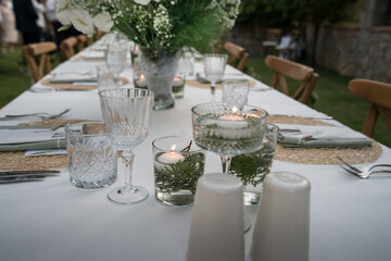 Beautiful decoration of a wedding banquet in the green garden in white colors. Serving the table with white tablecloth, candle, plates, glasses, cutlery, napkins decoration, with white flowers in vase