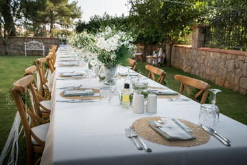 Beautiful decoration of a wedding banquet in the green garden in white colors. Serving the table with white tablecloth, plates, glasses, cutlery, napkins decoration, with white flowers in vase