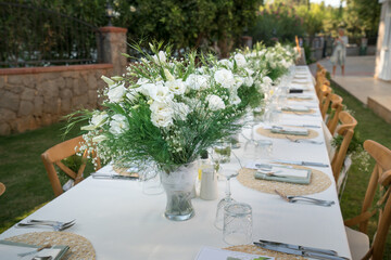 Beautiful decoration of a wedding banquet in the green garden in white colors. Serving the table with white tablecloth, plates, glasses, cutlery, napkins decoration, with white flowers in vase