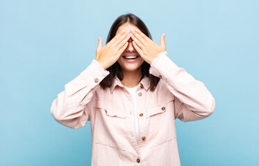 young pretty woman smiling and feeling happy, covering eyes with both hands and waiting for...