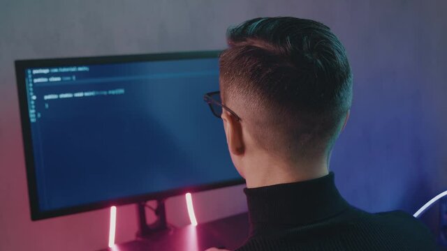Rear view of man programmer coding network security software indoors in neon lights. Hacker typing code on computer screen in dark office at night