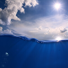 Fototapeta na wymiar Aquatic Marine Scenery With Bright sun on the sky. White clouds. Ocean Wave divided by waterline. Air bubbles and sun rays underwater