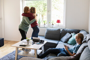 Smiling mother embracing daughter while son using phone at home