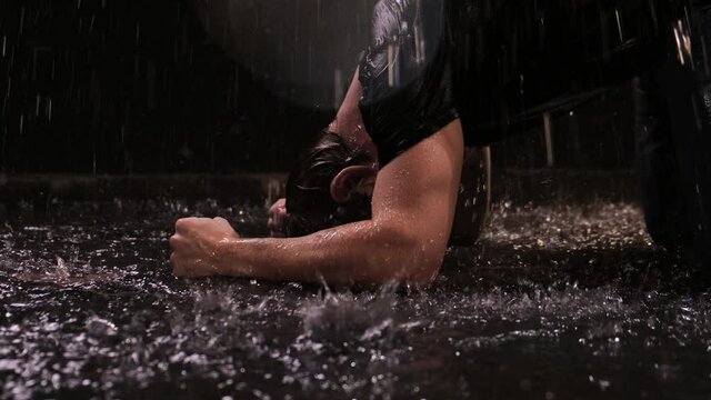 Young guy with long hair stands in the rain at night on the street in a black T-shirt. He is upset and falls to the ground and hits her with his hands. The concept of friend leaving life or big loss.