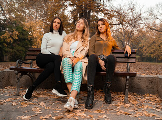 Three young adult, trendy woman, sitting on park bench in fall season