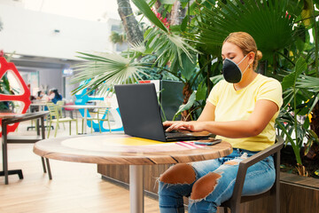Woman wearing a mask working at her desk looking at the laptop outdoors in a shopping center back...