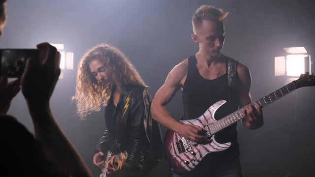 Fan takes phone pictures of two rock stars playing their guitars on stage surrounded by smoke. There is a lot of light. man in black T-shirt and woman in leather jacket play electric guitars