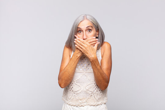 gray haired woman covering mouth with hands with a shocked, surprised expression, keeping a secret or saying oops