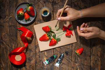 Gift card with live strawberries. A brush in female hands. Girl creates a postcard