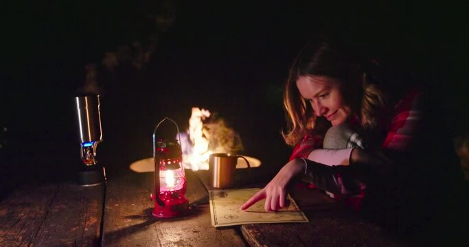 Woman on camping. Young female model in red classic shirt sitting at wooden table by campfire looking on the map and drinking hot tea enjoying outdoor adventure. Red lantern lightning the night scene