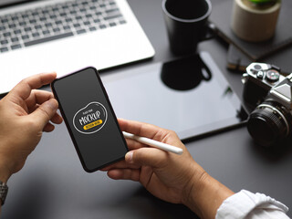 Male worker using mock up smartphone while working with laptop and camera on black table