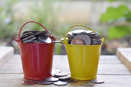 Many coins are in red and yellow buckets on a wooden table and have a blurred background.