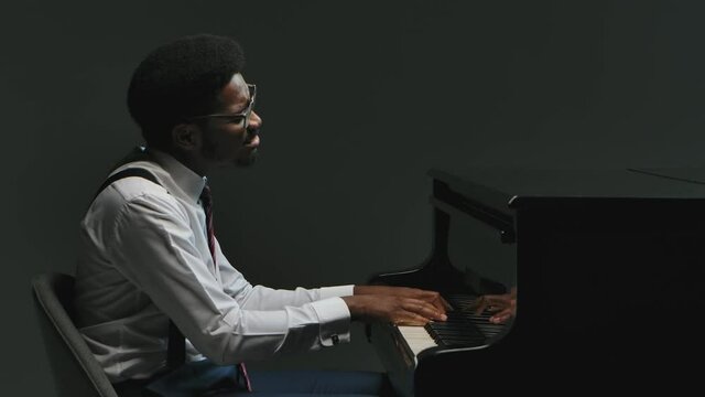 Happy young African American male playing the grand piano. A musician in an elegant suit and glasses touches his hands to black and white keys on a gray background. Close up. Slow motion.