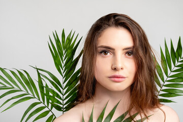 Fototapeta premium Natural skincare. Spa therapy. Portrait of sensual woman with perfect skin bare shoulder in green exotic palm leaves isolated on light empty space background. Organic cosmetology.