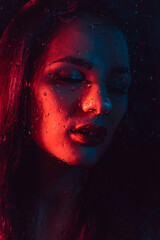 Portrait of sad young girl with red blue lighting behind glass with raindrops