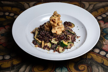 Traditional Spanish seafood chicken meat greens paella with zucchini and cheese on a dark wooden table. Fried shrimps, squids. Vegan, vegetarian. Black brown rice. Healthy food. White plate. Diet. 