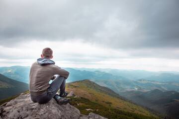 young strong man sitting on the rock at the top of the mountains peak