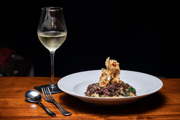 Traditional Spanish seafood chicken paella with zucchini on a dark wooden table. Fried shrimps, squids. Vegan, vegetarian. Black brown rice. Healthy food. Glass white wine. White plate. Diet.