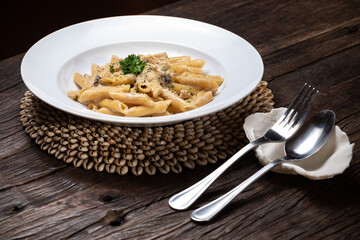 Homemade delicious penne pasta macaroni carbonara with white mushrooms chicken meet porcini in creamy cheese sauce in the white plate on the dark brown wood table. Parsley, spicy. Yellow, green.