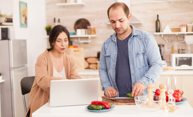 Couple prepares healthy salad looking at online recipe on laptop in kitchen. Man helping woman to prepare healthy organic dinner, cooking together. Romantic cheerful love relationship