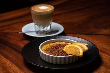 Creme brulee, burnt cream with orange with coffee, cappuccino on the dark wood table. French vanilla cream dessert with caramelised sugar on top on the white plate. Brown, orange, yellow, white. 