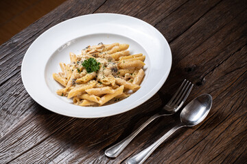 Homemade delicious penne pasta macaroni carbonara with white mushrooms chicken meet porcini in creamy cheese sauce in the white plate on the dark brown wood table. Parsley, spicy. Yellow, green.