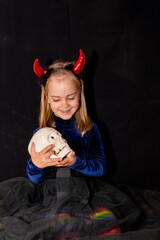  Little witch in a huge hat on her head on a black background. The girl is holding a silver skull....