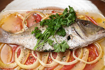 Baked fish with tomato, onion, sauteed vegetables. Whole sea fish and vegetables ingredients. Delicious marinated grilled or oven baked fish, parley, lemon on a white plate. Red, yellow, green.