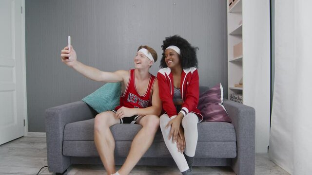 Caucasian man and african american woman retro style models relaxes on couch and takes photos on phone after work out session