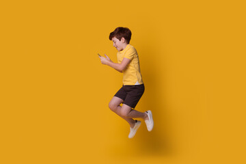 Fototapeta na wymiar Ginger caucasian boy wearing a yellow t-shirt is jumping on studio wall while chatting on mobile